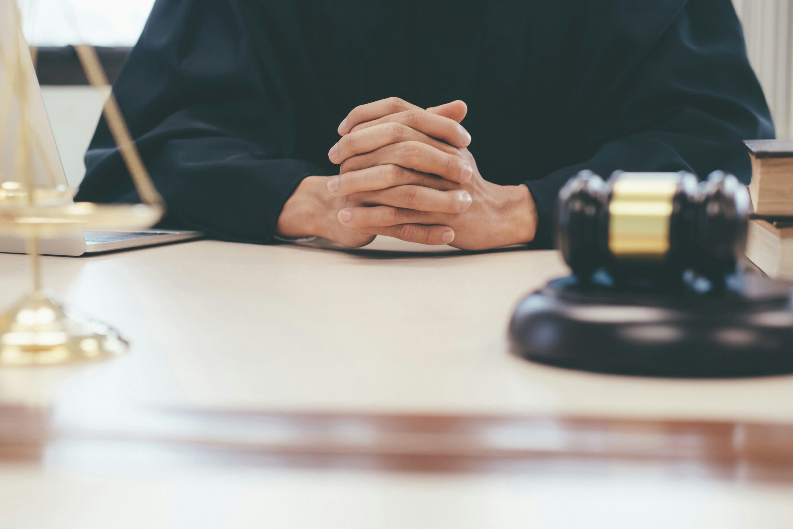 A judge sits at a desk with hands clasped, wearing a black robe. A gavel, a legal scale, and books are visible on the desk, suggesting a courtroom setting.