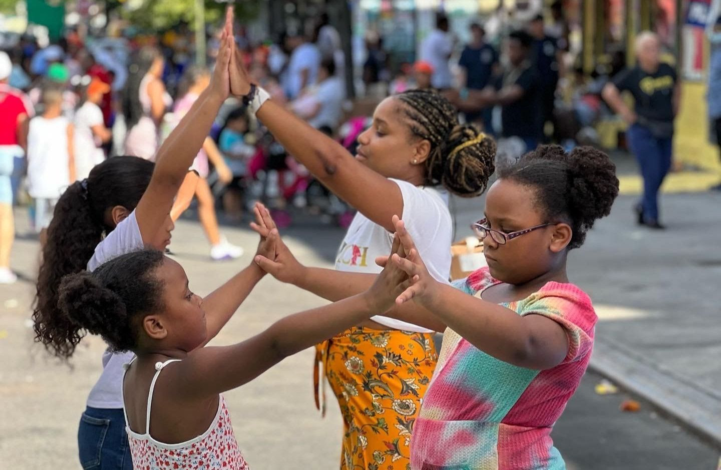 Four girls stand in pairs, facing each other and playing a hand-clapping game outdoors on a sunny day, with a crowd of people and colorful surroundings in the background.