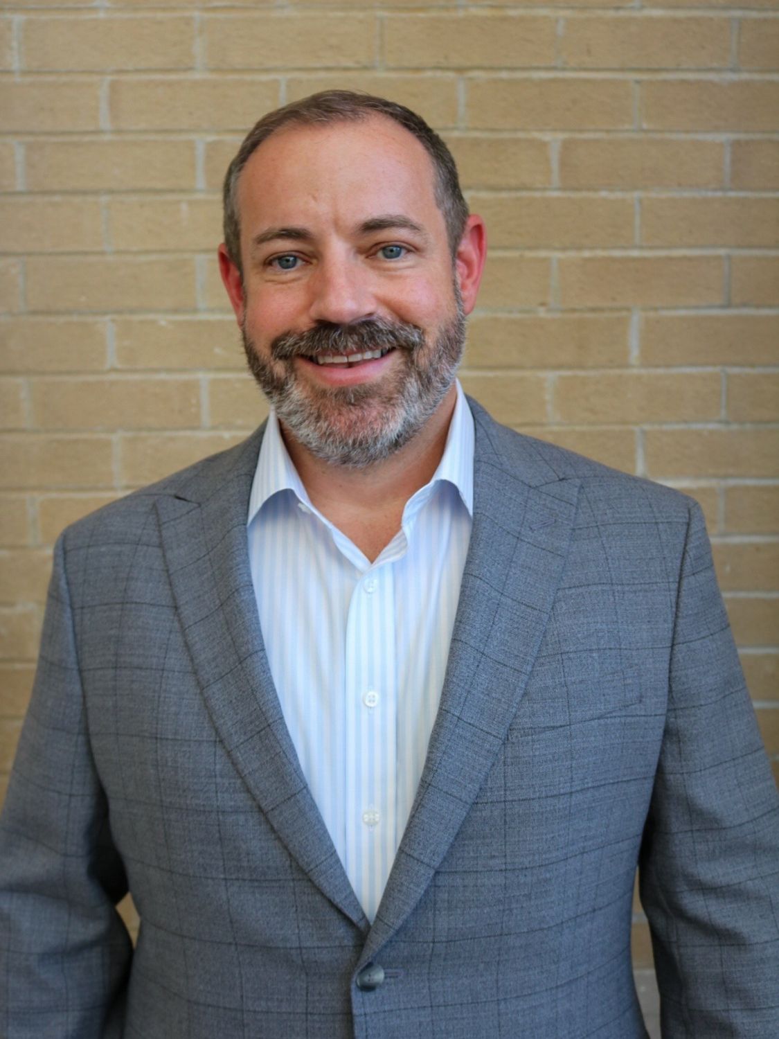 A man with short hair and a beard, wearing a gray checked suit jacket over a light blue dress shirt, stands smiling in front of a light brick wall.