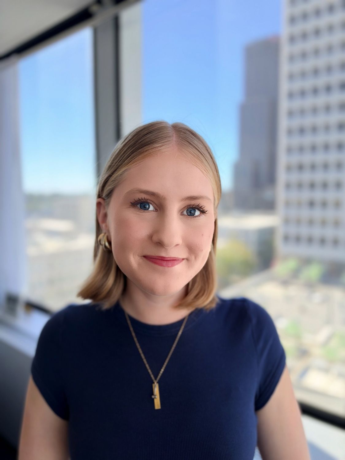A young woman with blonde hair, wearing a blue shirt and a gold necklace, smiles in front of a city office window.