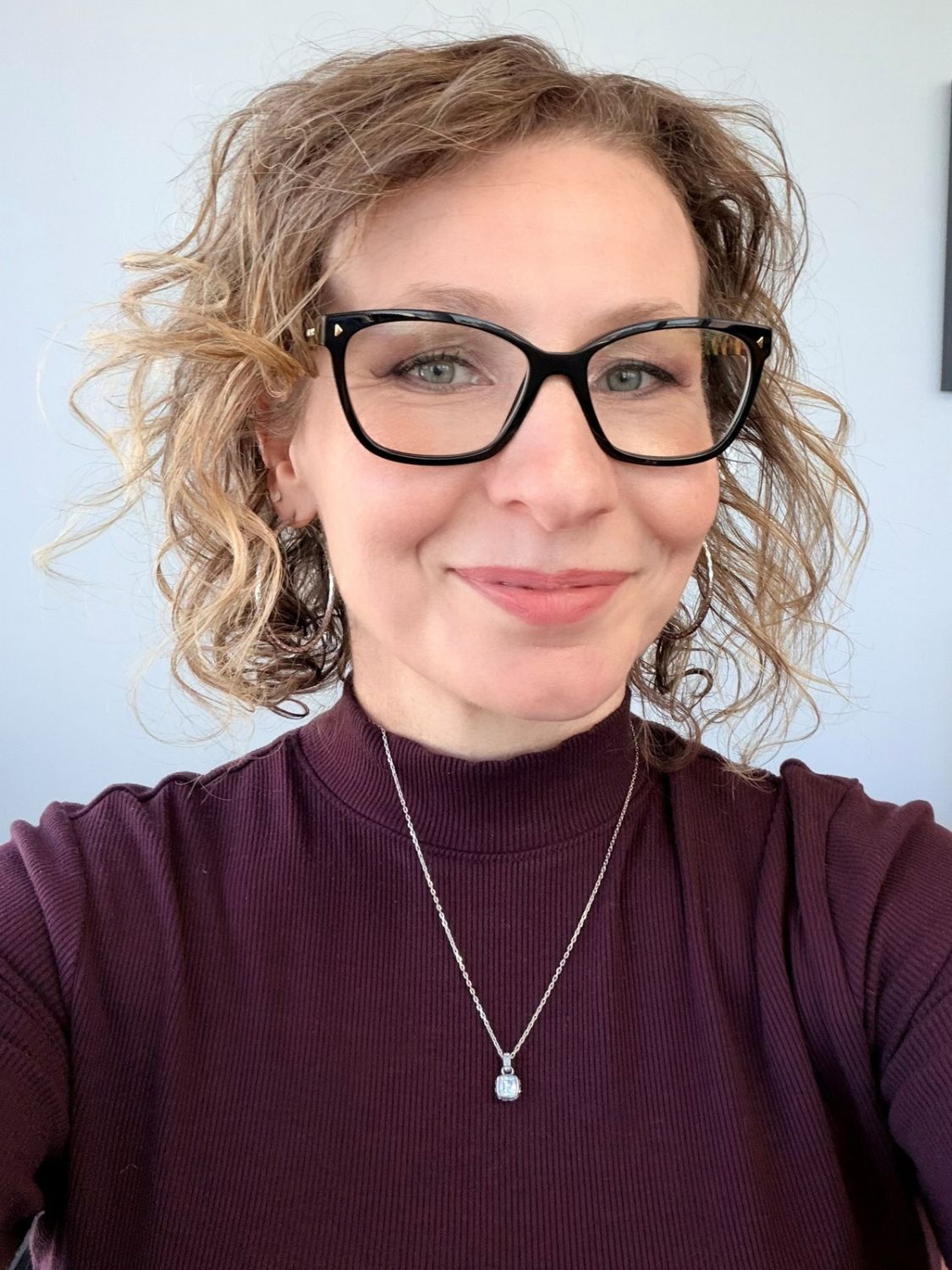 A woman with shoulder-length curly hair wearing black-rimmed glasses and a dark red mock-neck top with a silver necklace.