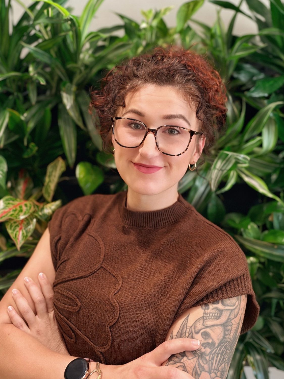 A woman with curly reddish-brown hair and glasses smiles with her arms crossed in front of a wall of green plants.